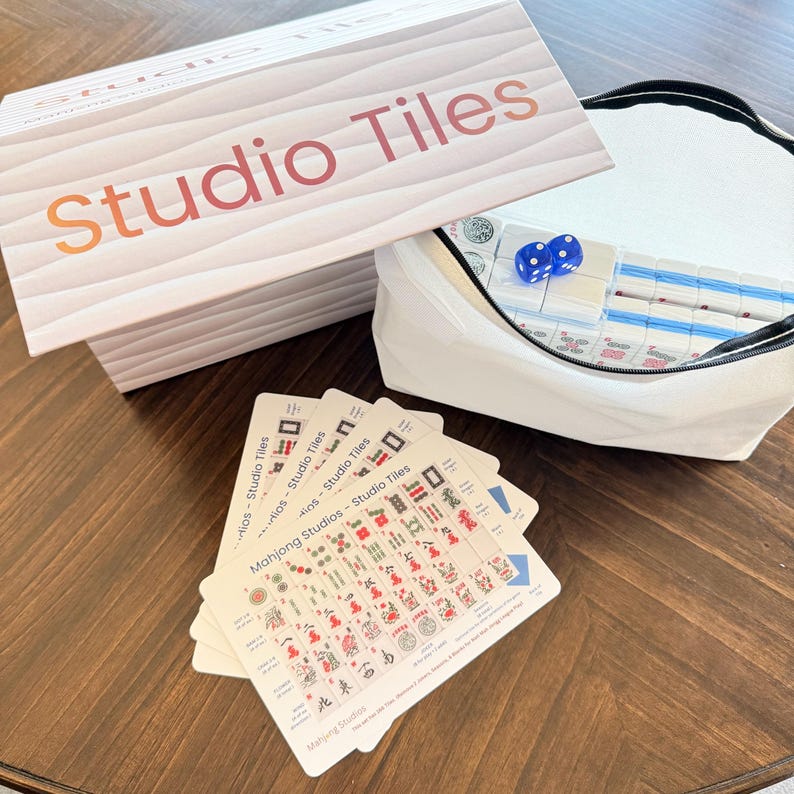 May include: A Studio Tiles Mahjong set with a wooden-look box and a white canvas bag. The box and bag are open, revealing tiles, dice, and cards. The cards display colorful symbols and text. The game is set on a dark wood surface.