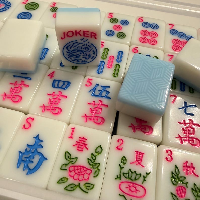 May include: A close-up of a Mahjong set with white tiles and colorful markings. The tiles feature Chinese characters, floral designs, and numbers. One tile reads "JOKER" with a dragon design. The set includes blue and pink accents.