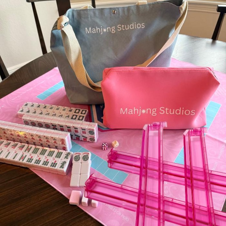 May include: A Mahjong game set with a light blue tote bag and a pink zippered pouch, both with "Mahjong Studios" printed on them. The set includes pink tile racks, dice, and several stacks of tiles, all arranged on a pink game mat.