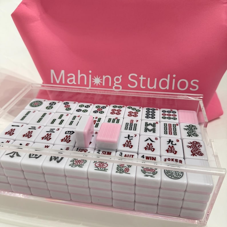 May include: A clear acrylic case containing a set of white Mahjong tiles with pink accents. The tiles feature Chinese characters and symbols. A pink bag with the text "Mahj*ng Studios" is in the background.