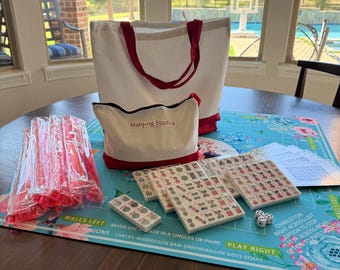 May include: A Mahjong game set with a white tote bag and a matching pouch, both with red accents. The set includes tiles, dice, and a game mat with floral and text details. The pouch reads "Mahjong Studios".