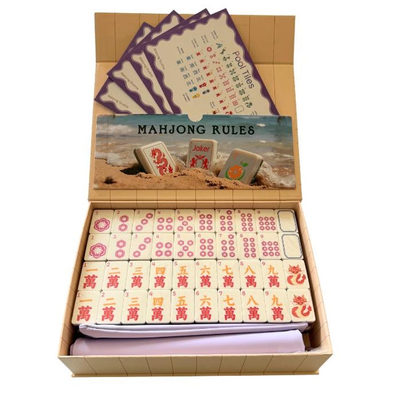 May include: A beige box containing a Mahjong set with white tiles featuring red and black Chinese characters and symbols. The set includes a rule booklet with a beach scene, a reference card for pool tiles, and a folded light purple fabric cloth.