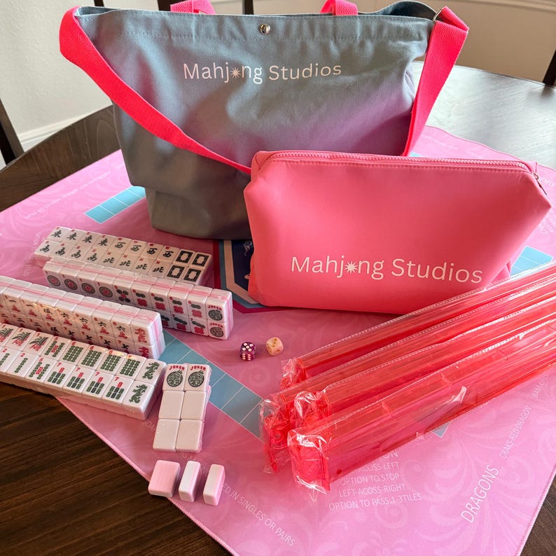 May include: A Mahjong set in pink and grey. The set includes tiles, dice, a pink zipped pouch and a grey tote bag, both with "Mahjong Studios" printed on them. The game is set on a pink mat. The set also includes red plastic sticks.