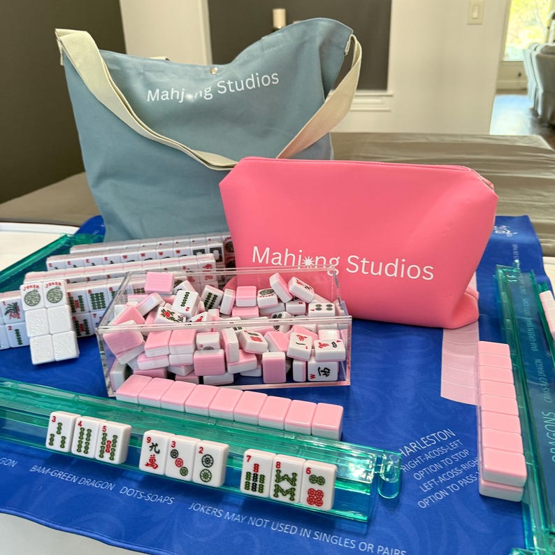 May include: A Mahjong set featuring pink and white tiles, a blue tote bag, and a pink cosmetic bag, all branded with the "Mahjong Studios" logo. The set includes tile racks and a blue playing mat. The text "CHARLESTON" is visible on the mat.