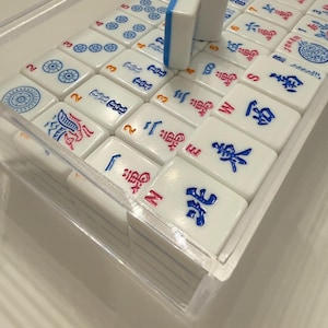 Puede incluir: Un juego de Mahjong completo con fichas blancas con s&iacute;mbolos y n&uacute;meros azules, rojos y negros. Las fichas est&aacute;n cuidadosamente dispuestas en una caja de pl&aacute;stico transparente.