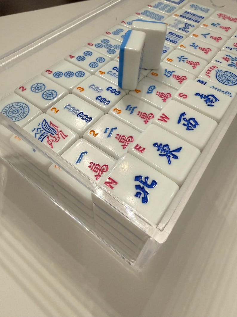 Puede incluir: Un juego de Mahjong completo con fichas blancas con s&iacute;mbolos y n&uacute;meros azules, rojos y negros. Las fichas est&aacute;n cuidadosamente dispuestas en una caja de pl&aacute;stico transparente.