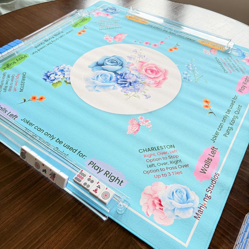 May include: A light blue Mahjong game mat with floral and butterfly designs. The mat features a central circular design with blue and pink roses. Clear plastic tile racks are visible, along with Mahjong tiles.