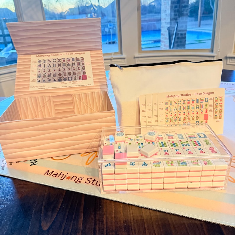 May include: A Mahjong set with pink and white tiles in a clear acrylic case. The set includes a wooden box, a canvas bag, and a printed guide. The text "Mahjong Studios - Rose Dragon" is visible.
