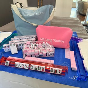 May include: A Mahjong set with pink and white tiles, a clear acrylic tile holder, and a red tile rack. The set includes a blue tote bag and a pink pouch, both with the text "Mahjong Studios". The set is on a blue mat.