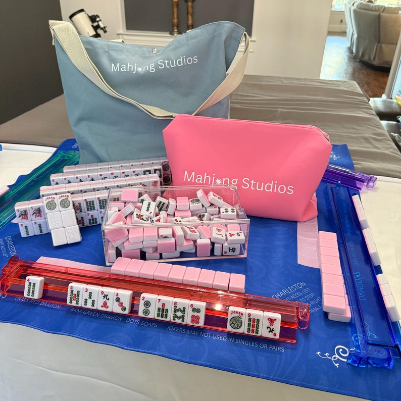 May include: A Mahjong set with pink and white tiles, a clear acrylic tile holder, and a red tile rack. The set includes a blue tote bag and a pink pouch, both with the text "Mahjong Studios". The set is on a blue mat.