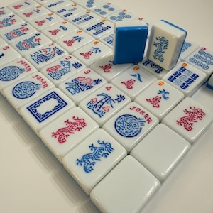 Puede incluir: Un juego de fichas de Mahjong blancas con s&iacute;mbolos y caracteres azules y rojos. Dos fichas est&aacute;n de pie, una azul y otra blanca con un dise&ntilde;o de drag&oacute;n azul. Las fichas est&aacute;n dispuestas sobre una superficie blanca.