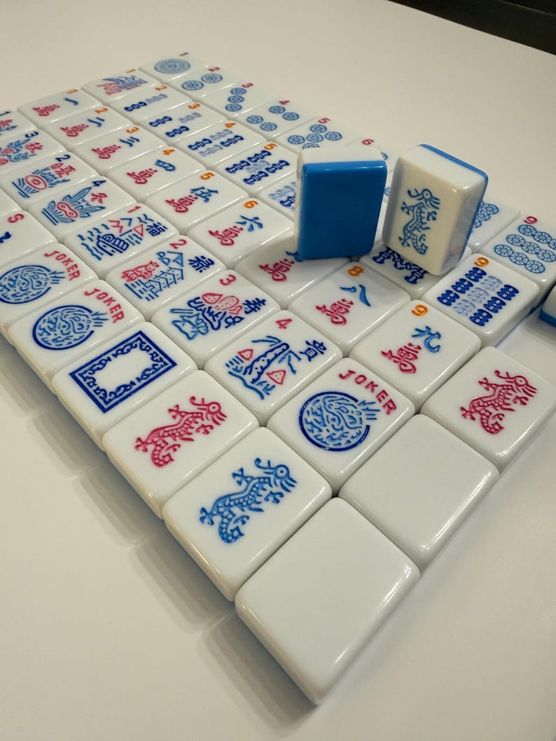 Puede incluir: Un juego de fichas de Mahjong blancas con s&iacute;mbolos y caracteres azules y rojos. Dos fichas est&aacute;n de pie, una azul y otra blanca con un dise&ntilde;o de drag&oacute;n azul. Las fichas est&aacute;n dispuestas sobre una superficie blanca.