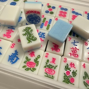 May include: A close-up of a Mahjong set with white tiles. Some tiles have blue, green, and pink symbols and characters. One tile reads "JOKER" with a blue dragon design. Other tiles have numbers and suit symbols.