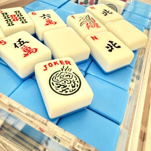 May include: A close-up of a Mahjong set with ivory-colored tiles and blue foam padding. The tiles display Chinese characters and symbols in red and black ink. One tile reads "JOKER" with a green dragon design. The set is in a clear acrylic case.