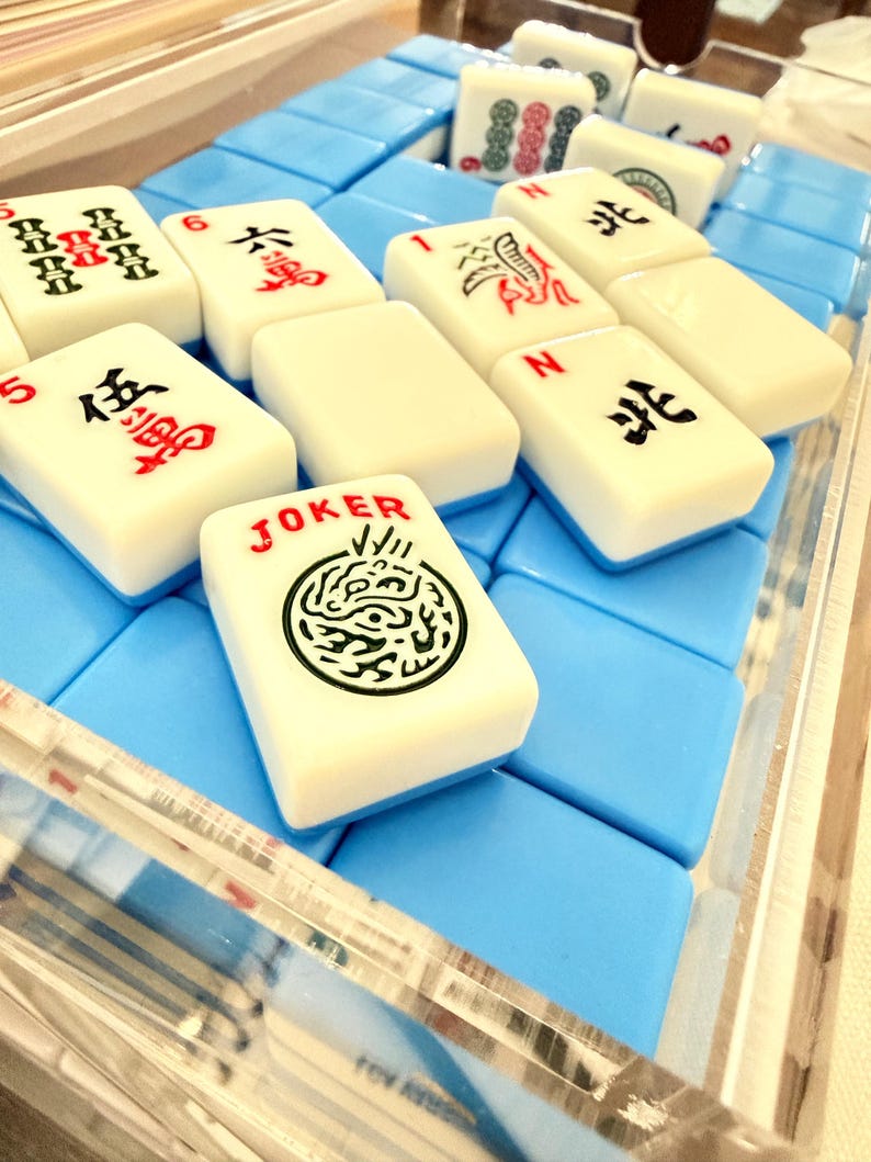 May include: A close-up of a Mahjong set with ivory-colored tiles and blue foam padding. The tiles display Chinese characters and symbols in red and black ink. One tile reads "JOKER" with a green dragon design. The set is in a clear acrylic case.