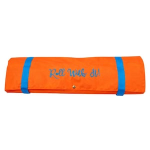 Puede incluir: Una esterilla naranja enrollada, asegurada con dos correas azules. La esterilla tiene el texto bordado "Roll With It!" en escritura azul. Se ve un cierre de botón a presión dorado.