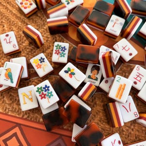 May include: A collection of Mahjong tiles, featuring a mix of white, brown, and red colors. Some tiles display floral designs, numbers, and the word "Joker". The tiles are scattered, suggesting a game in progress.