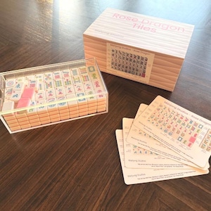 May include: A Rose Dragon Tiles mahjong set. The set includes a clear acrylic case filled with tiles, a wooden box with the text "Rose Dragon Tiles", and several reference cards. The tiles are cream-colored with various symbols and characters.