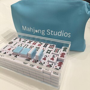 May include: A complete Mahjong set in a clear acrylic case, with white tiles featuring red and green symbols. The set includes blue and white dice and is accompanied by a light blue zippered pouch with the text "Mahjong Studios."