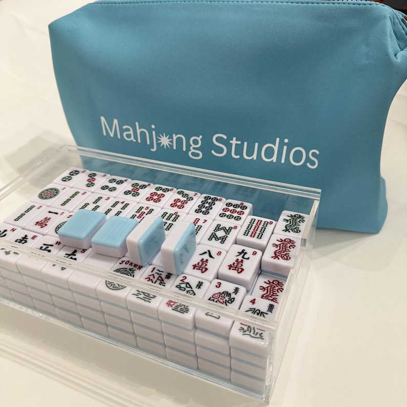 May include: A complete Mahjong set in a clear acrylic case, with white tiles featuring red and green symbols. The set includes blue and white dice and is accompanied by a light blue zippered pouch with the text "Mahjong Studios."