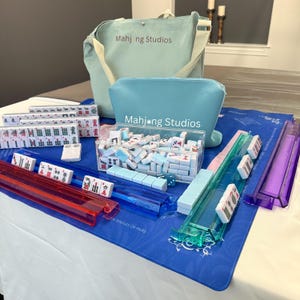 May include: A complete Mahjong set with tiles, racks, and accessories, displayed on a blue mat. The set includes a clear acrylic box filled with tiles, and a light blue tote bag and pouch with the text "Mahj*ng Studios".