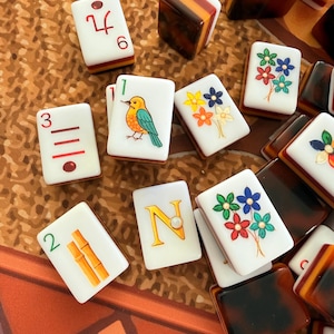 May include: A collection of white Mahjong tiles with various designs and numbers. Some tiles feature illustrations of a bird, bamboo stalks, and colorful flowers. Other tiles display the number 6, the number 3, and the letter N.