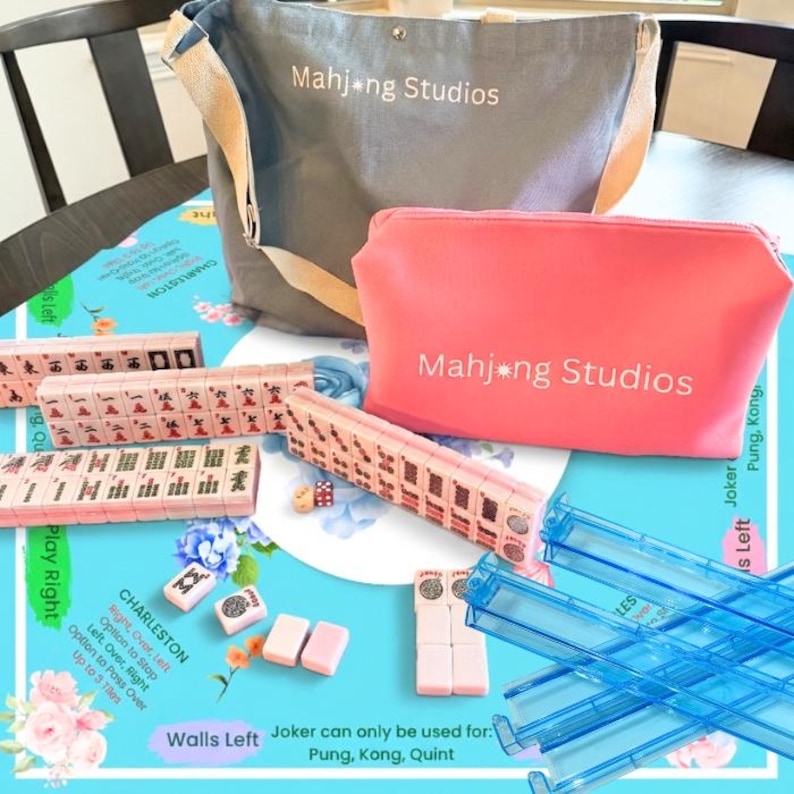 May include: A Mahjong set with a gray tote bag and a pink zippered pouch, both with "Mahjong Studios" printed on them. The set includes pink tiles, dice, and blue tile racks, all arranged on a blue floral patterned surface.