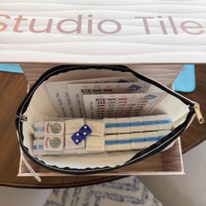 May include: A Mahjong set with white tiles and blue accents, including dice and instruction cards, is displayed inside a zippered pouch. The pouch is inside a box with the text "Studio Tiles".