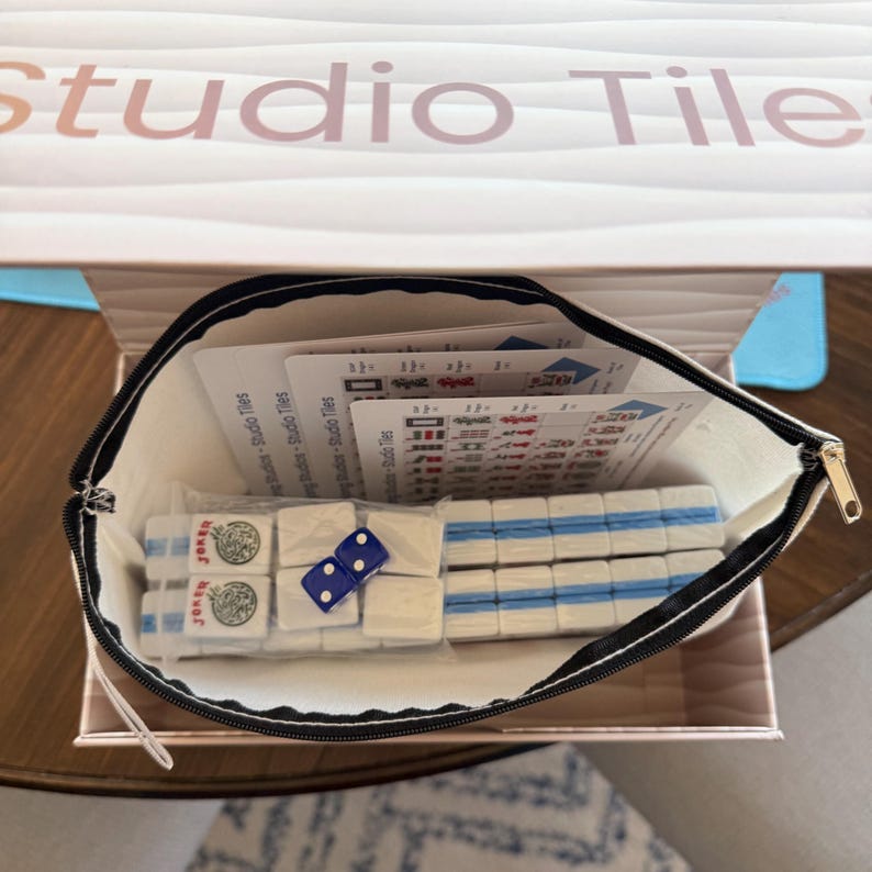 May include: A Mahjong set with white tiles and blue accents, including dice and instruction cards, is displayed inside a zippered pouch. The pouch is inside a box with the text "Studio Tiles".