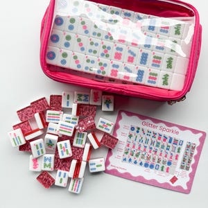 May include: A pink zippered case containing a set of white Mahjong tiles with colorful symbols. Some tiles are scattered outside the case. A pink card with the text "Glitter Sparkle" and Mahjong instructions is also visible.