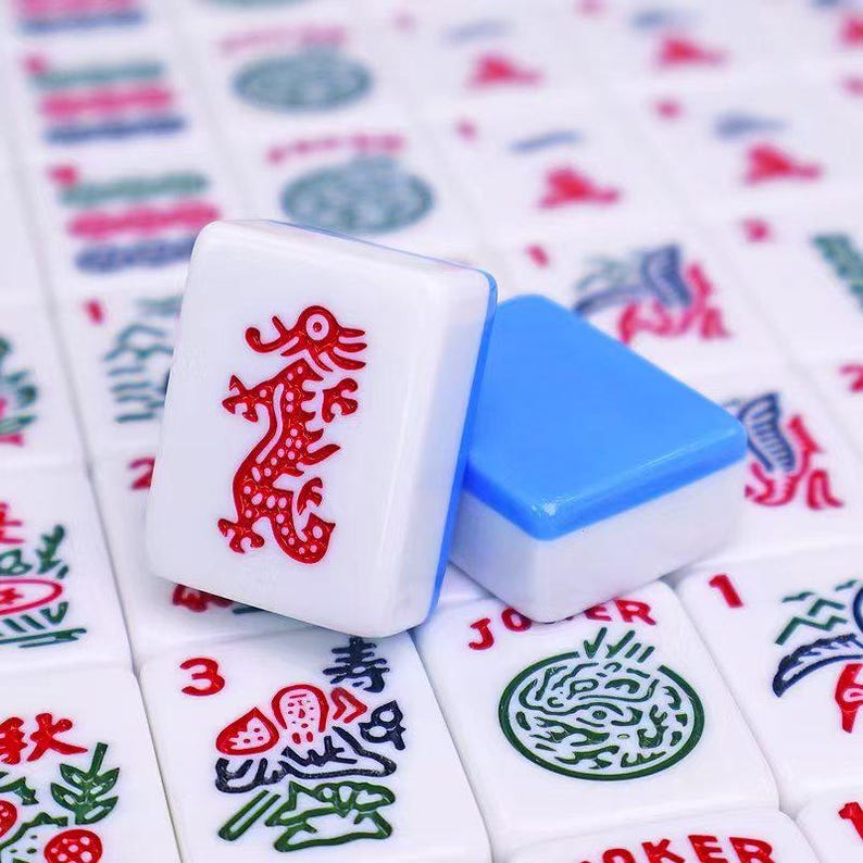 May include: Close-up of Mahjong tiles. One tile features a red dragon design on a white background. Another tile is blue on top and white on the bottom. Other tiles are visible in the background.