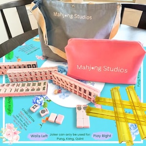 May include: A complete Mahjong set with tiles, dice, and racks, along with a gray tote bag and a pink cosmetic bag, all bearing the "Mahjong Studios" logo. The set is arranged on a blue floral patterned surface.