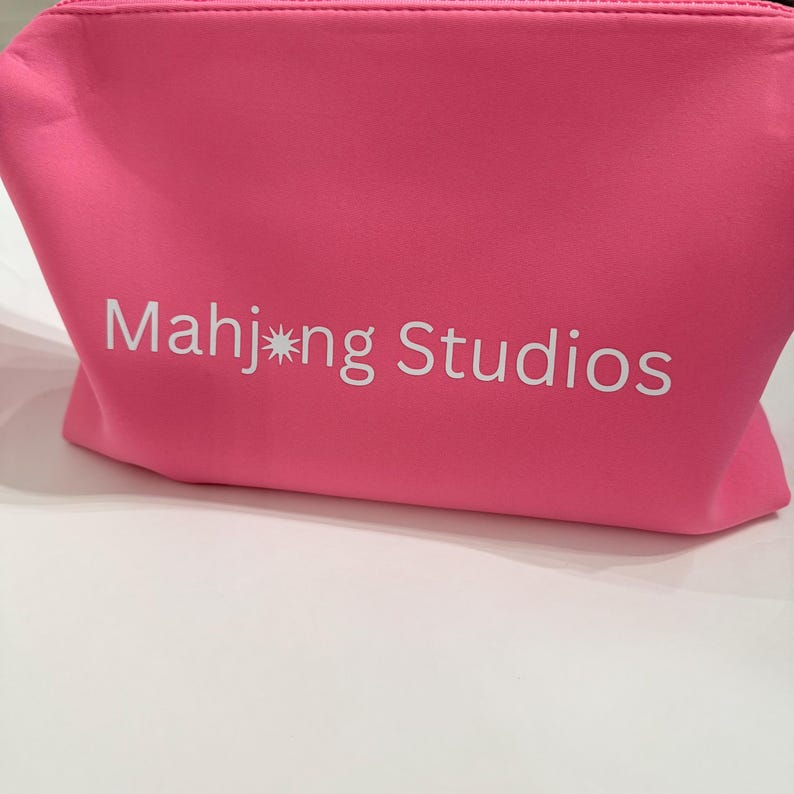 May include: A vibrant pink cosmetic bag featuring the white text "Mahj*ng Studios". The bag is made of a soft material and has a zip closure. It is placed on a white surface.