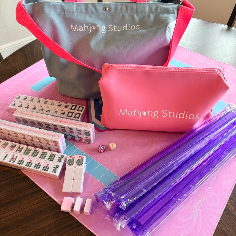 May include: A Mahjong set featuring a pink mat, tiles, dice, and accessories. The set includes a grey tote bag and a pink zipped pouch, both with "Mahjong Studios" printed on them. Purple tile racks are also included.