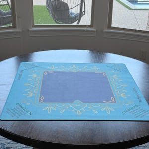 May include: A square, light blue mat with a central purple rectangle and decorative gold-colored floral designs. The word "CHARLESTON" is printed in white on the mat, along with other text. The mat is on a dark brown table.