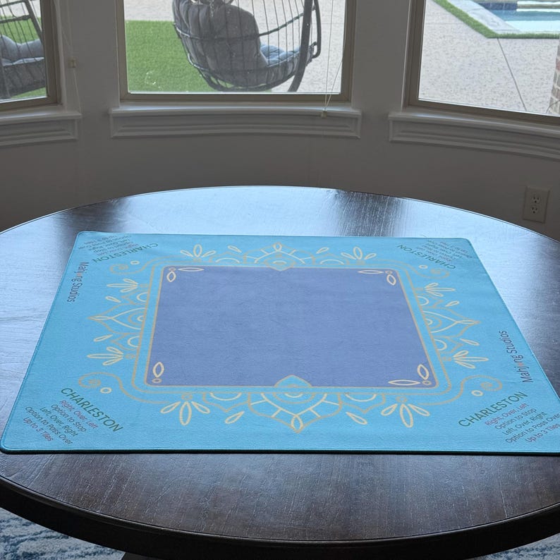 May include: A square, light blue mat with a central purple rectangle and decorative gold-colored floral designs. The word "CHARLESTON" is printed in white on the mat, along with other text. The mat is on a dark brown table.