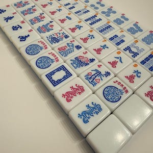 Puede incluir: Un conjunto de fichas de Mahjong blancas dispuestas sobre una superficie. Las fichas presentan s&iacute;mbolos y caracteres rojos y azules. Algunas fichas tienen la palabra "JOKER". Las piezas del juego son cuadradas y tienen bordes redondeados.