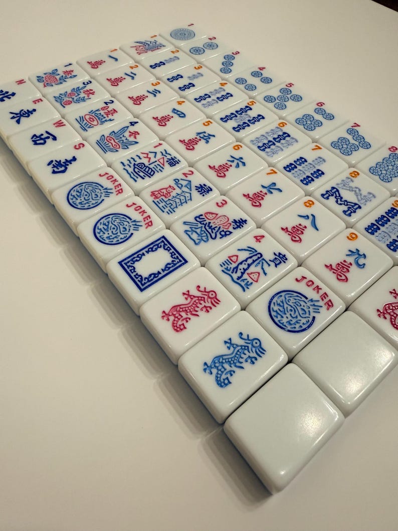 Puede incluir: Un conjunto de fichas de Mahjong blancas dispuestas sobre una superficie. Las fichas presentan s&iacute;mbolos y caracteres rojos y azules. Algunas fichas tienen la palabra "JOKER". Las piezas del juego son cuadradas y tienen bordes redondeados.
