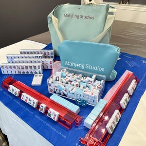 May include: A complete Mahjong set with tiles, racks, and accessories. The set includes a blue tote bag and a matching zippered pouch, both with the text "Mahj ng Studios". The tiles are white with red, green, and blue markings.