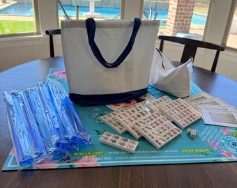 May include: A white tote bag with navy blue trim and handles sits on a floral game board. Several wrapped blue game pieces, game tiles, and dice are also visible. The game board has text that reads "WALLS LEFT NEVER USE..." and "PLAY RIGHT".