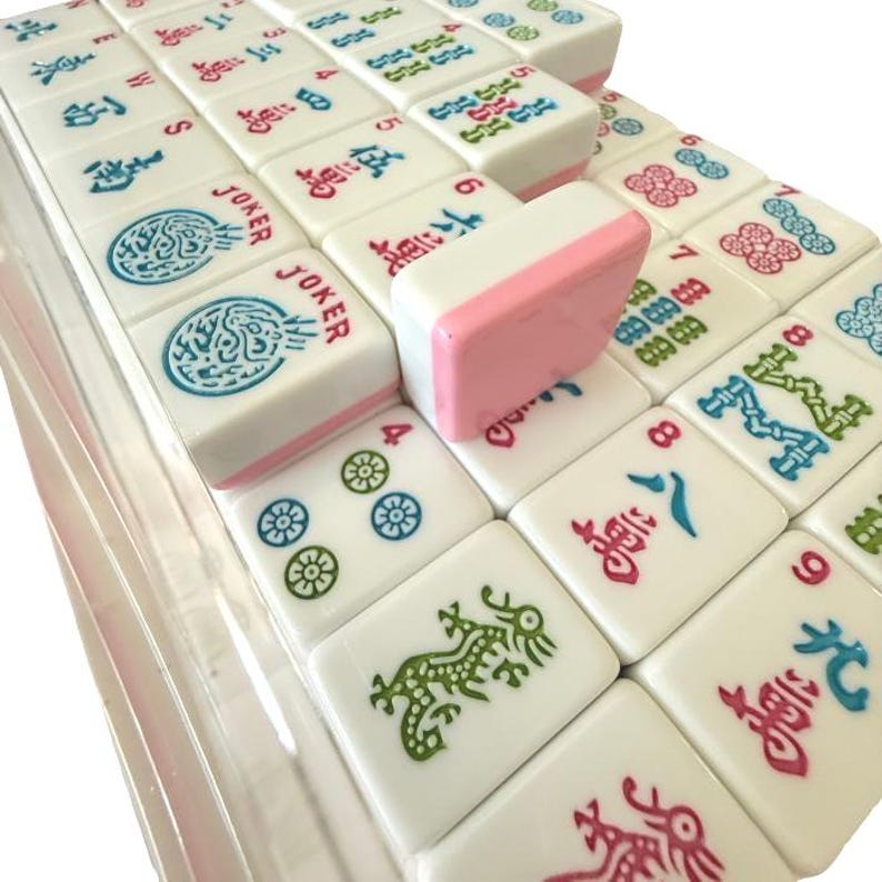 May include: A set of white Mahjong tiles with colorful symbols and characters. The tiles have pink edges and are arranged in a clear plastic case. The game pieces feature blue, green, and red designs.