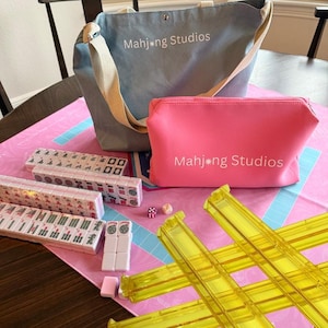 May include: A Mahjong set with a blue tote bag and a pink cosmetic bag, both with "Mahjong Studios" printed on them. The set includes tiles, dice, and yellow tile racks, all arranged on a pink mat.