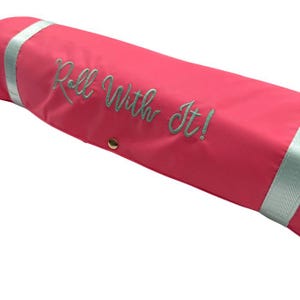 May include: A bright pink, cylindrical bag with the embroidered phrase "Roll With It!" in a cursive font. The bag has light blue accents and a gold-colored snap closure.