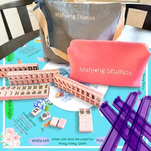 May include: A complete Mahjong set with tiles, dice, and racks, along with a gray tote bag and a pink cosmetic bag, all bearing the "Mahjong Studios" logo. The set is arranged on a blue floral patterned surface.