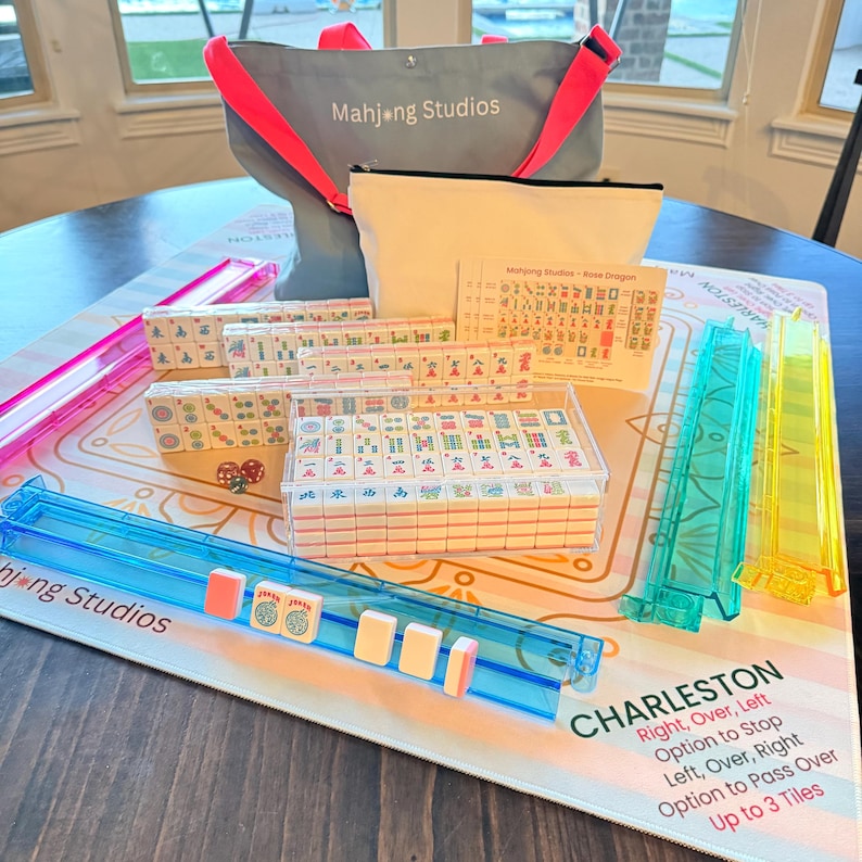 May include: A complete Mahjong set with tiles, racks, and a game mat. The set includes a gray tote bag with the text "Mahjong Studios", a beige pouch, and a game guide. The game mat has the word "Charleston" printed on it.
