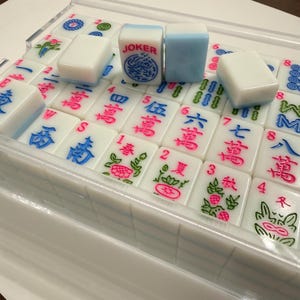 May include: A set of white Mahjong tiles with blue and pink markings, displayed in a clear plastic case. The tiles feature Chinese characters and symbols, including a "JOKER" tile with a blue dragon design. The game set is designed for recreational use.