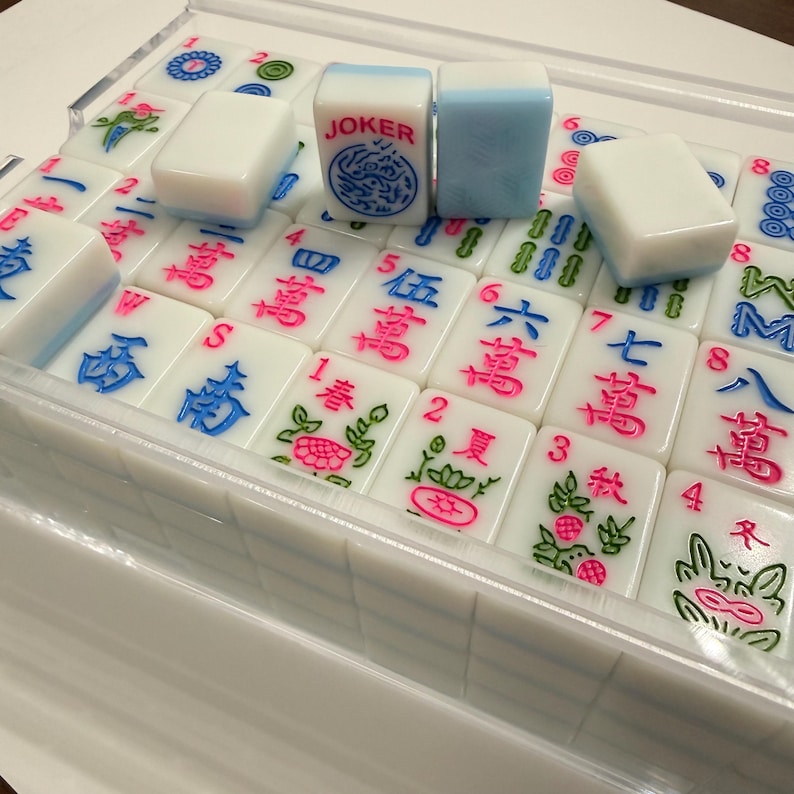 May include: A set of white Mahjong tiles with blue and pink markings, displayed in a clear plastic case. The tiles feature Chinese characters and symbols, including a "JOKER" tile with a blue dragon design. The game set is designed for recreational use.