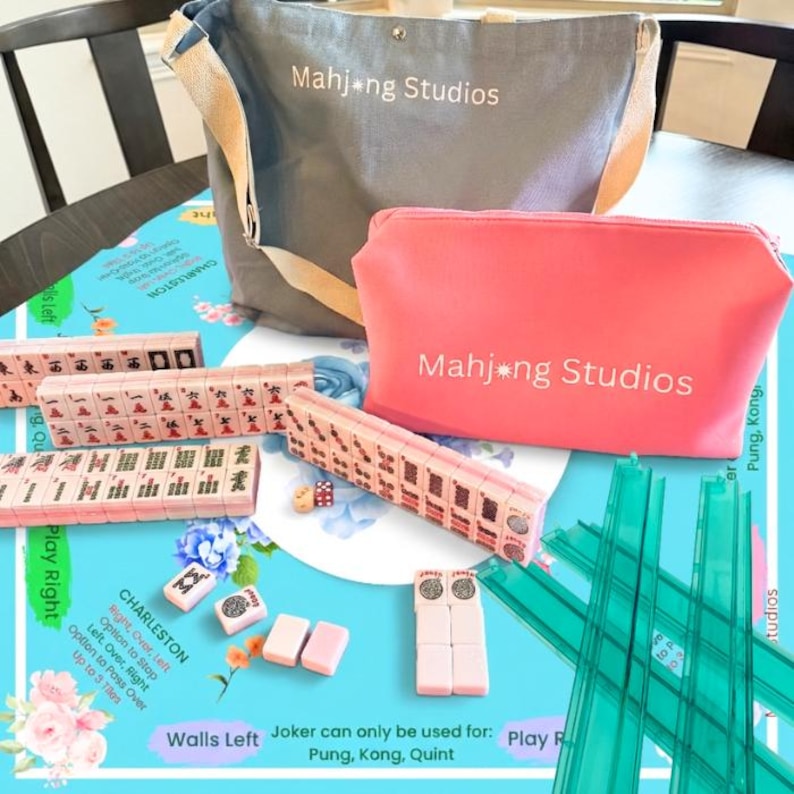May include: A Mahjong set with a gray tote bag and a pink cosmetic bag, both with the text "Mahjong Studios". The set includes pink tiles, dice, and green tile racks, all arranged on a blue floral patterned surface.