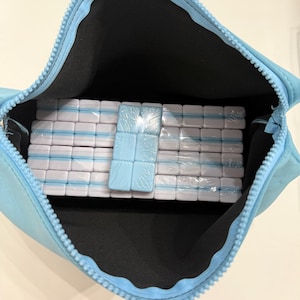 May include: A light blue zipped pouch filled with stacks of white and light blue rectangular tiles, wrapped in clear plastic. The pouch is open, revealing the contents inside. The tiles appear to be game pieces.