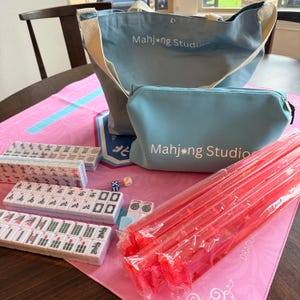 May include: A complete Mahjong set, including tiles, dice, and racks, with a light blue tote bag and pouch, both with "Mahjong Studio" printed on them. The set is arranged on a pink playing mat.